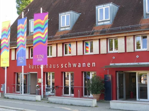 Rotes Gebäude mit mehreren Fenstern und bunten Fahnen, beschriftet 'kulturnausschwanen'. (KI generiert)