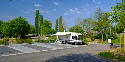 Wohnmobil auf einem Stellplatz mit Markise und Tisch, umgeben von Bäumen und blühenden Sträuchern.