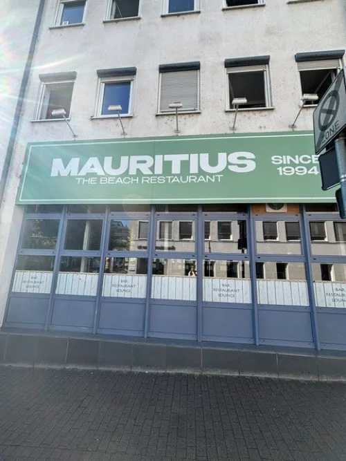 Fassade des Restaurants Mauritius seit 1994 mit blau-grüner Markise. (KI generiert)