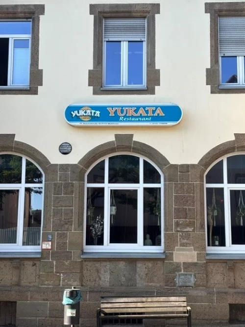 Fassade eines Yukata Restaurants mit blauem Schild und drei großen Fenstern. (KI generiert)