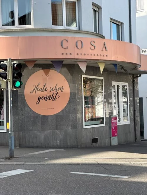 Ladenfront mit dem Namen COSA und einem Schild mit der Aufschrift Heute schon genäht? (KI generiert)