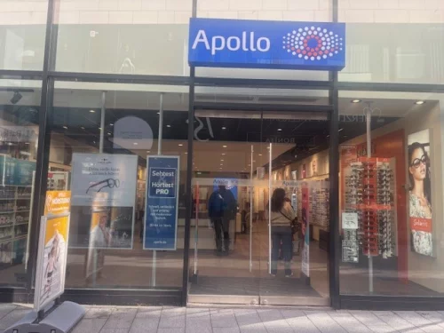 Apollo-Filiale mit zwei Personen vor dem Eingang, Schaufenster mit Werbung. (KI generiert)