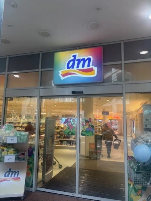 dm-Drogeriemarkt mit zwei Personen vor Regalen, beleuchtetes Logo über Eingang. (KI generiert)