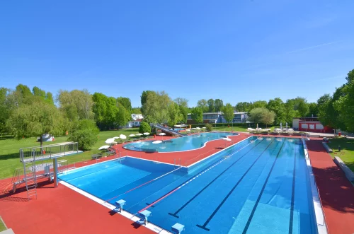 Großer Pool mit blauer Wasserfläche, umgeben von roten Liegewiesen und Bäumen im Hintergrund. (KI generiert)