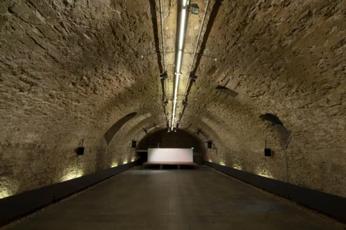 Ein langer, beleuchteter Tunnel mit Steinwänden und einem hellen Bildschirm am Ende. (KI generiert)
