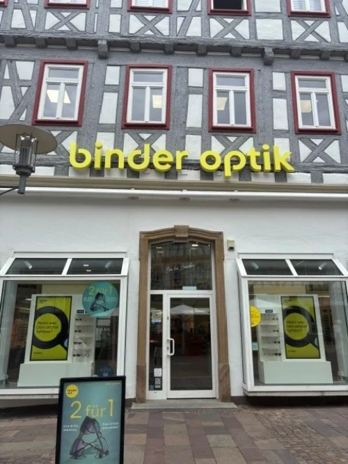 Fassade eines Optikgeschäfts mit Schaufenstern und gelb-grünen Werbeplakaten. (KI generiert)