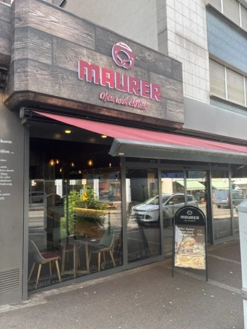 Fassade eines Cafés mit dem Namen 'Maurer' in roter Schrift, geöffnet. (KI generiert)