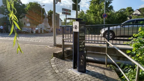 Ein Fahrradverleihstand mit zwei Fahrrädern und einer Verleihstation in einer Stadtumgebung mit gepflastertem Boden und einer Brücke. (KI generiert)