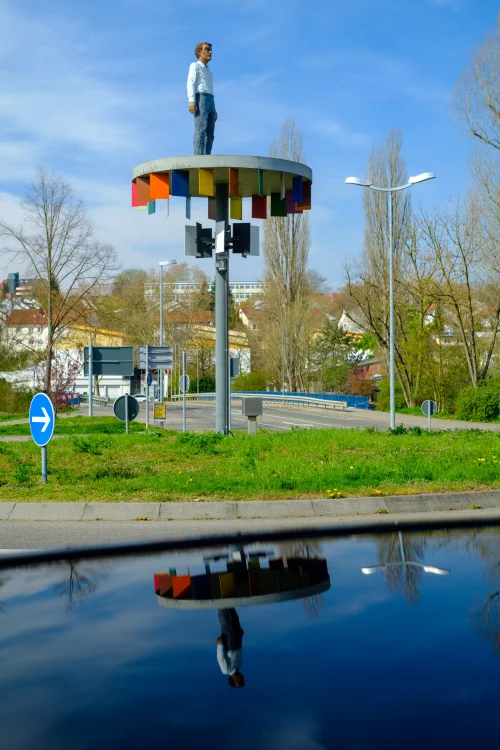 Eine Statue eines Mannes steht auf einer bunten, kreisförmigen Plattform auf einem hohen Pfahl. Die Plattform spiegelt sich im Wasser darunter. (KI generiert)