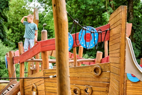 Kind spielt auf Holzspielplatz mit Klettergerüst und blauer Rutsche. (KI generiert)