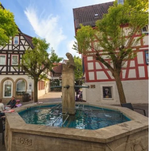 Ein Brunnen mit einer Statue in der Mitte, umgeben von historischen Fachwerkhäusern und Bäumen. Menschen sitzen auf Bänken. (KI generiert)